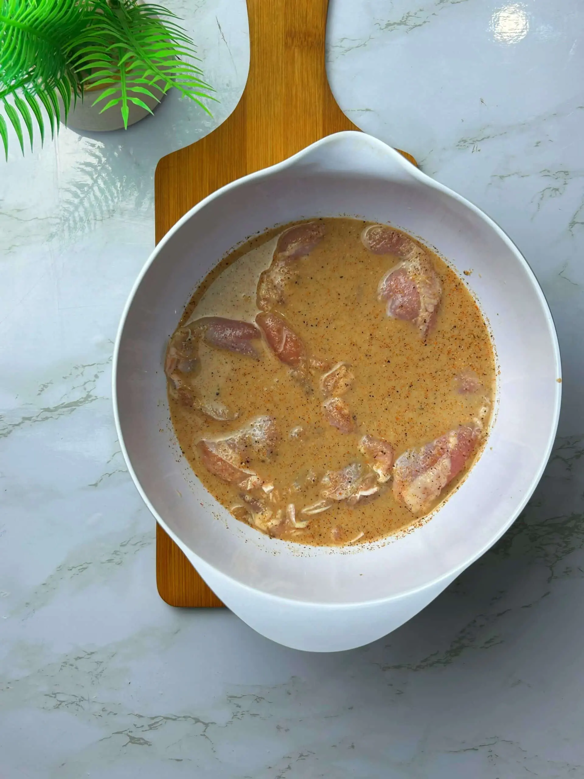 Boneless chicken thighs marinating in seasoned buttermilk brine in a white bowl on a wooden board